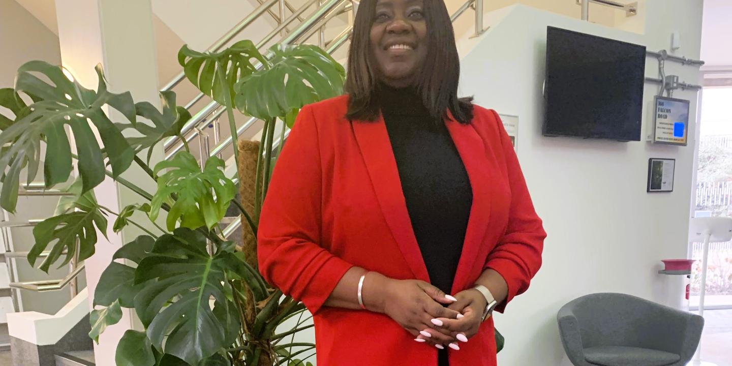 A woman wearing a red jacket stands formally beside an office stair case.