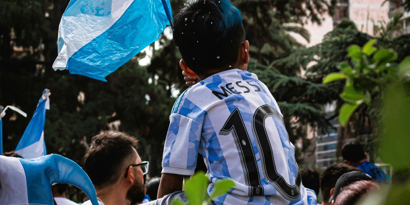 A child on the shoulders of a parent wears a light blue and white stripped football top, waves the Argentinian flag