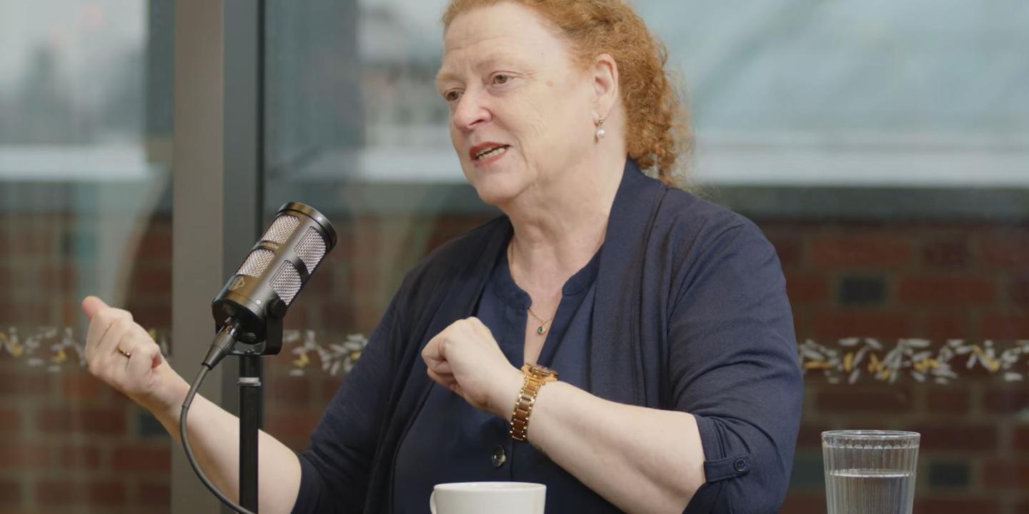 A woman guest on a podcast sits at a table with a mic in front, gesturing to the side with both hands.