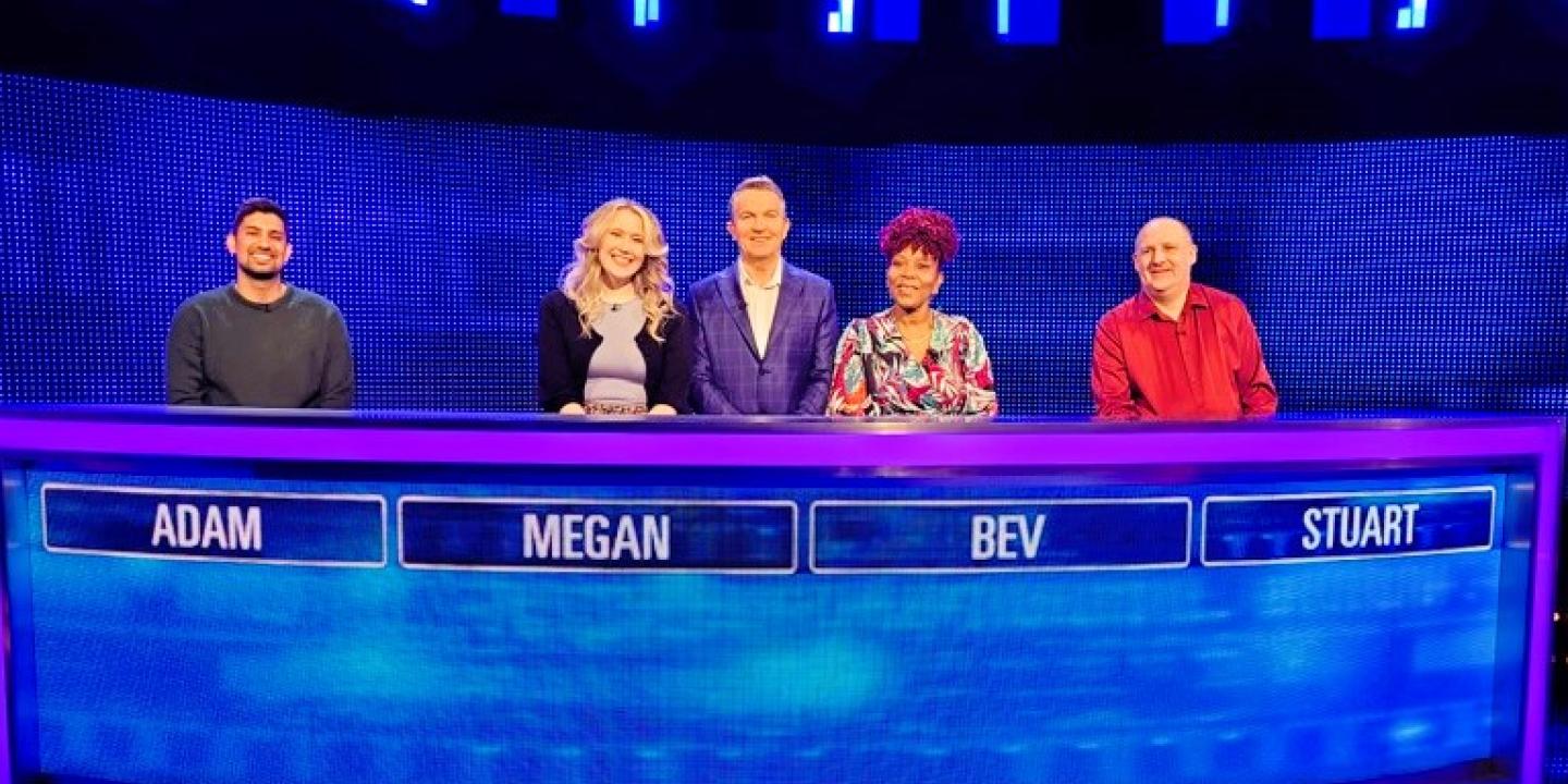 Four quiz show contestants stand behind a table with the show host.