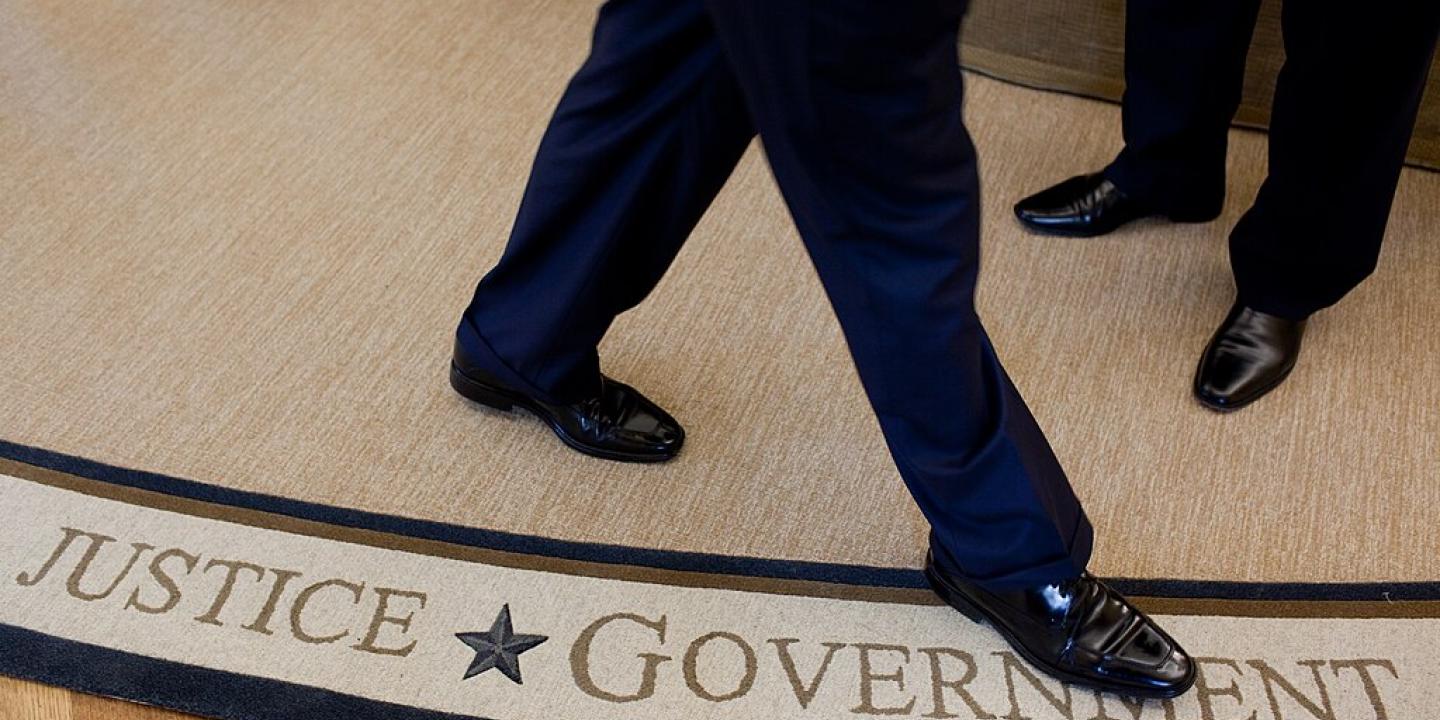 The feet and legs of someone walking on a white rug, beside the words Justice and Government woven in to it,