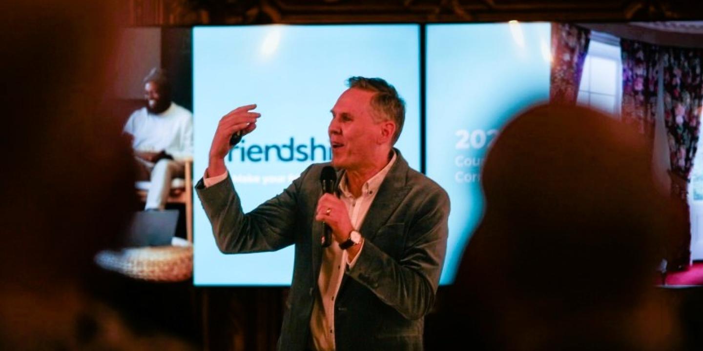 A speaker, standing in front of a screen, beckons with one hand, holding a mic with the other.