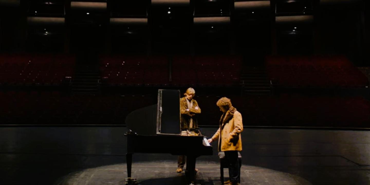Two people sit and stand next to a grand piano on a stage.