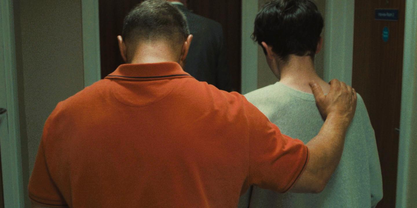 A father walks with his son away from the camera, his hand on his son's shoulder.