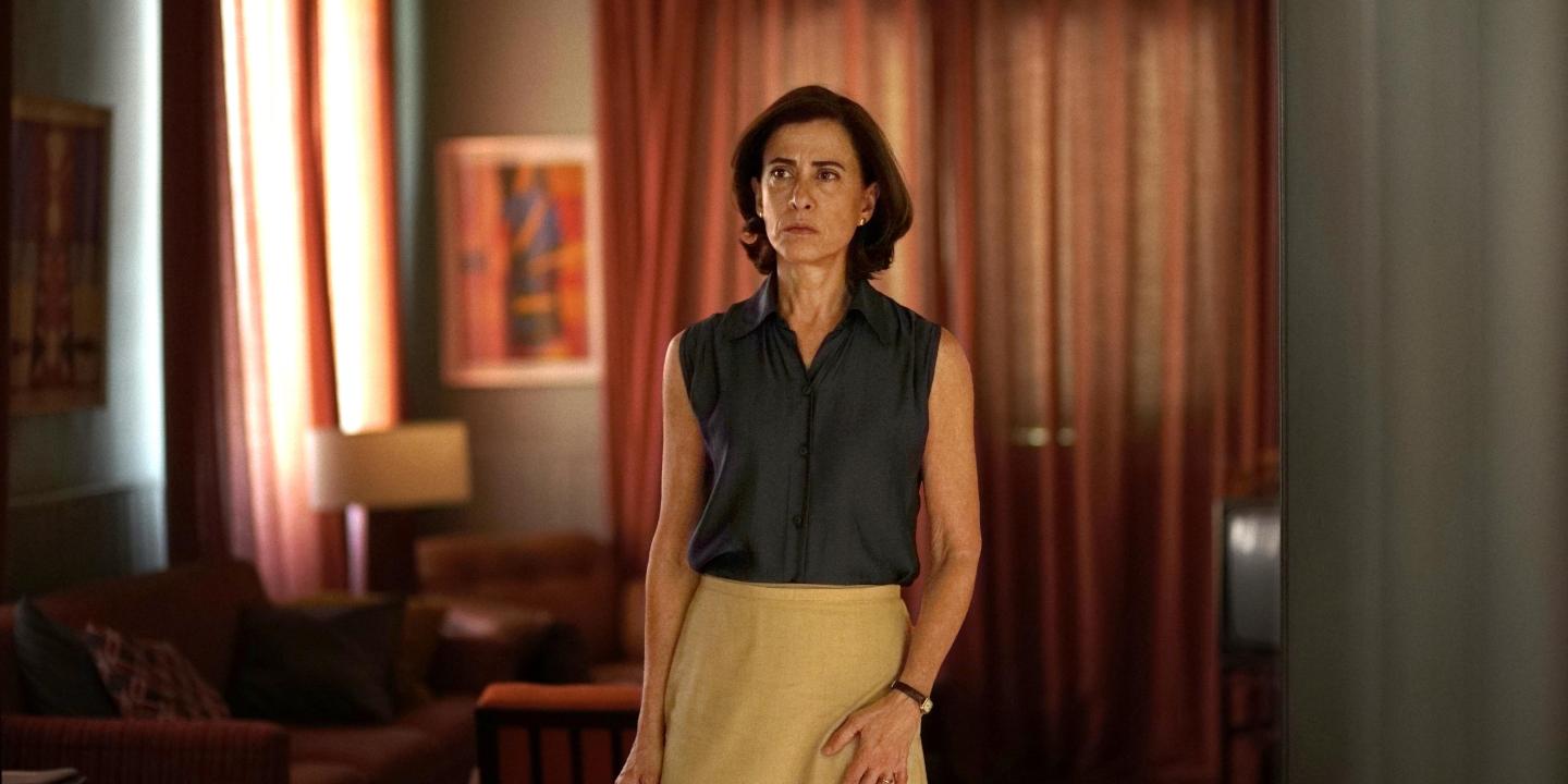 A woman, wearing 70s clothing, stands, looking apprehensive.