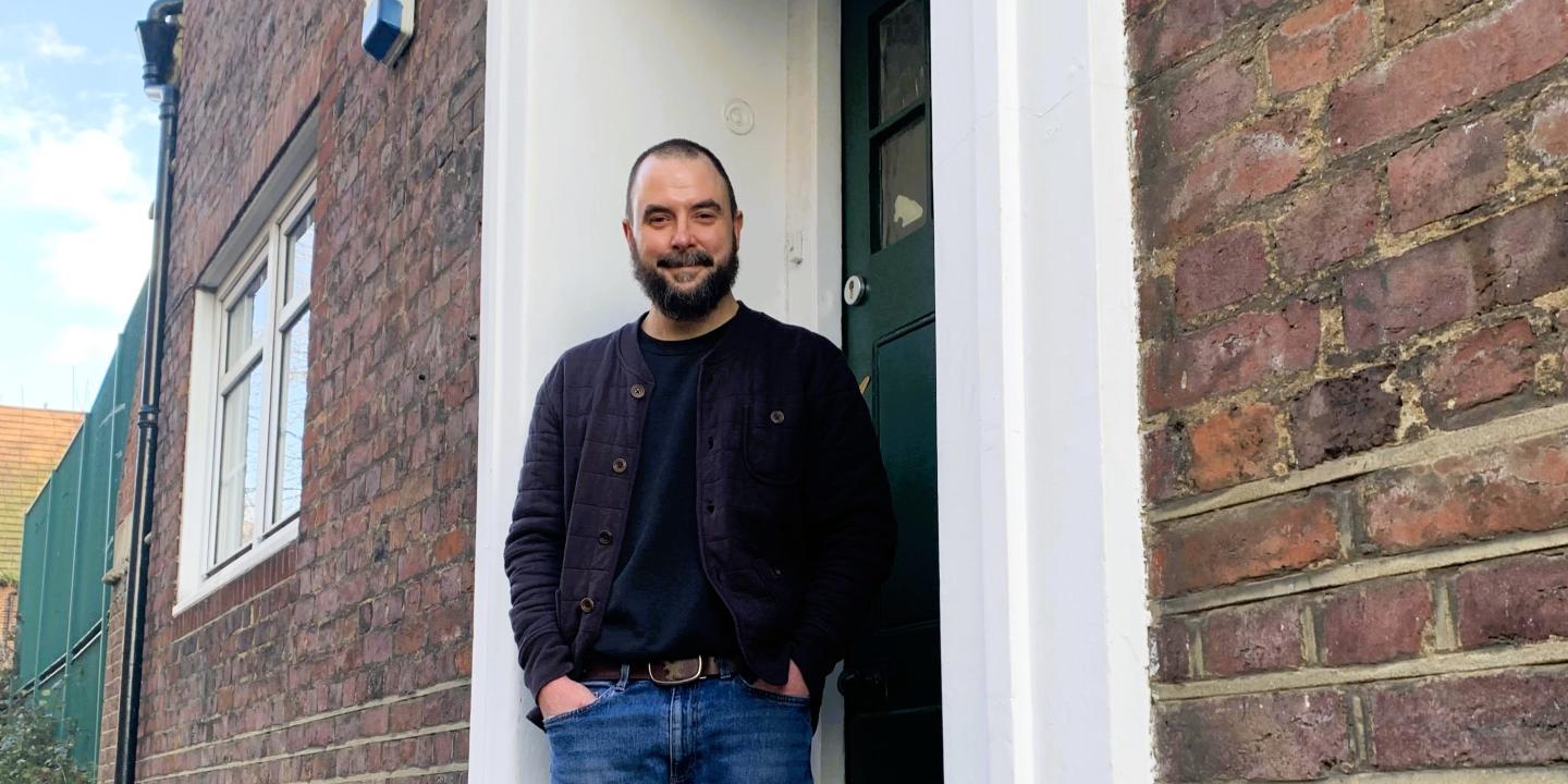 A man stands in a relaxed way on the doorstep of a brick building.