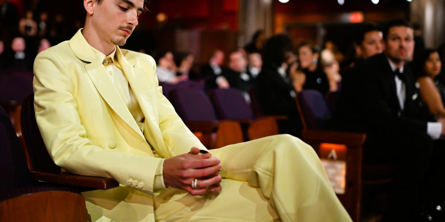 Timothee Chalamet, wearing light yellow suit reposes in a seat a the Oscars, clasping his hands, his eyes closed.