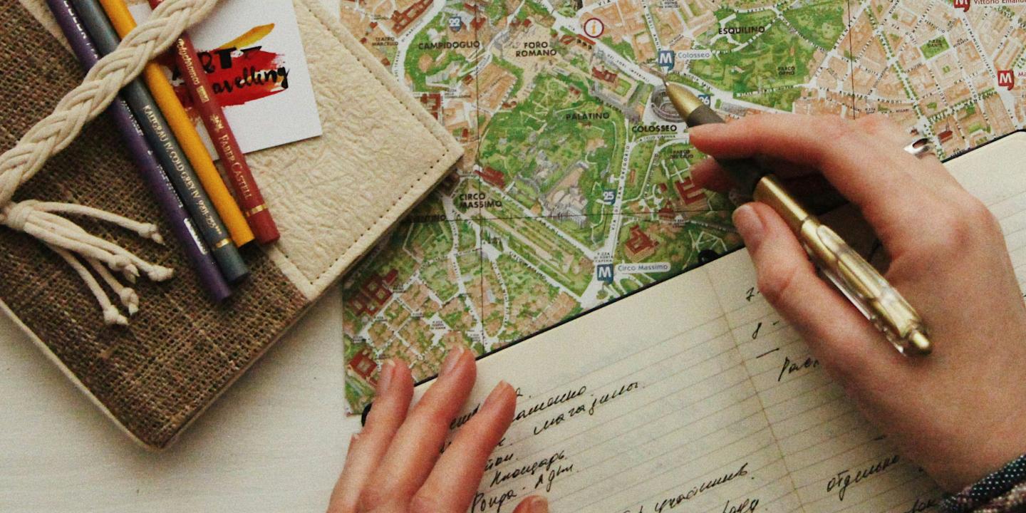 A hand holds a pen over a map, at the side is closed journal and colour pencils.