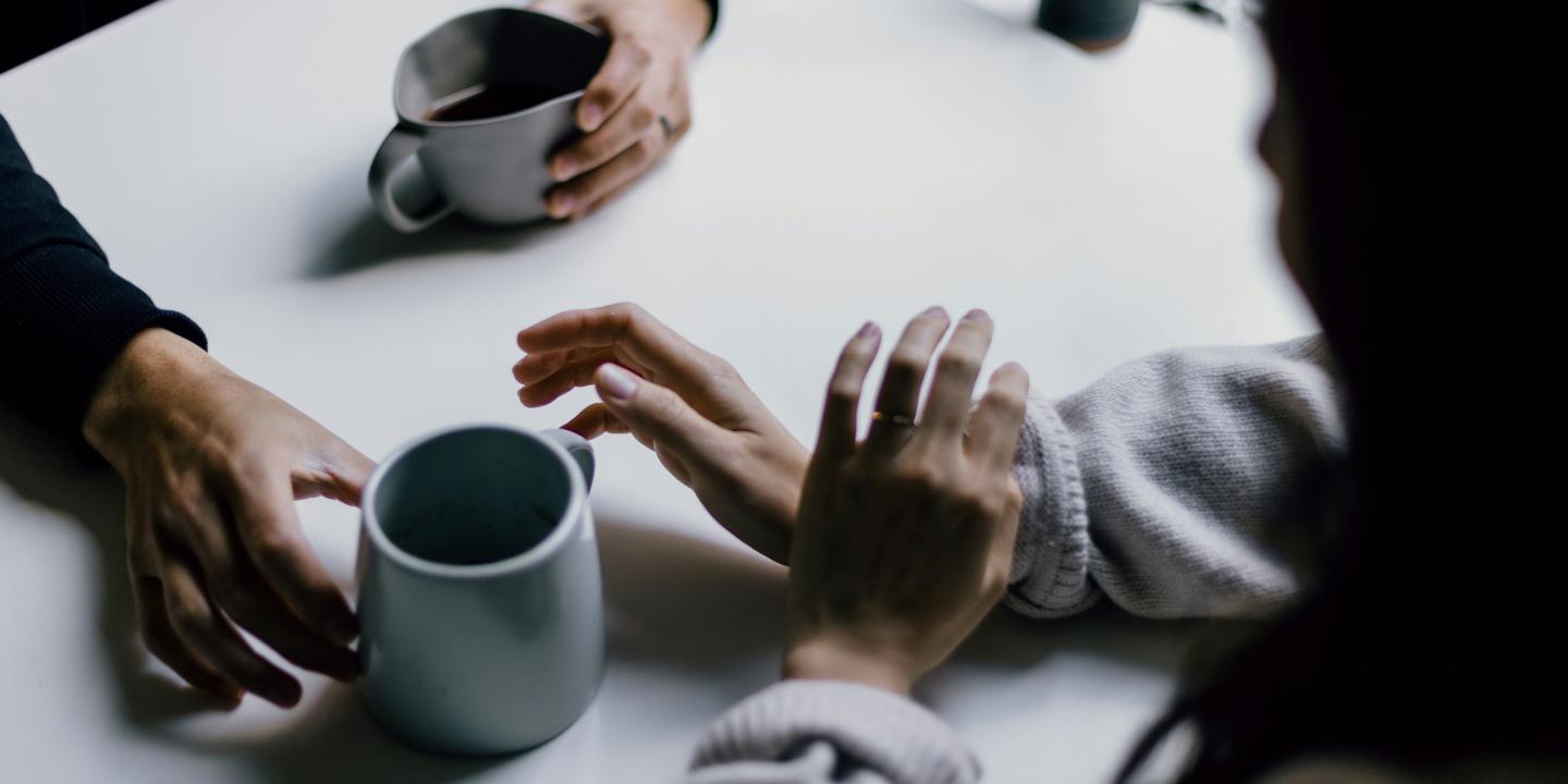A cup of coffee is offered across a table to a hand that is hesitant.