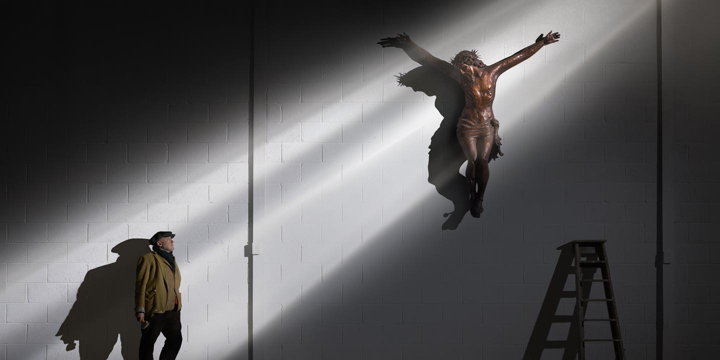 A man looks up a shaft of light that illuminates him and a crucifix higher up a wall.