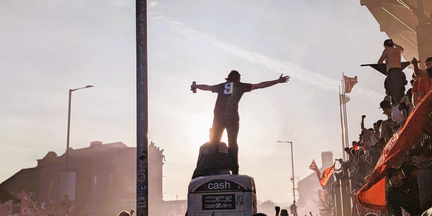 Amid celebrating football fans, one stands on top of a kiosk with outstretched arms.