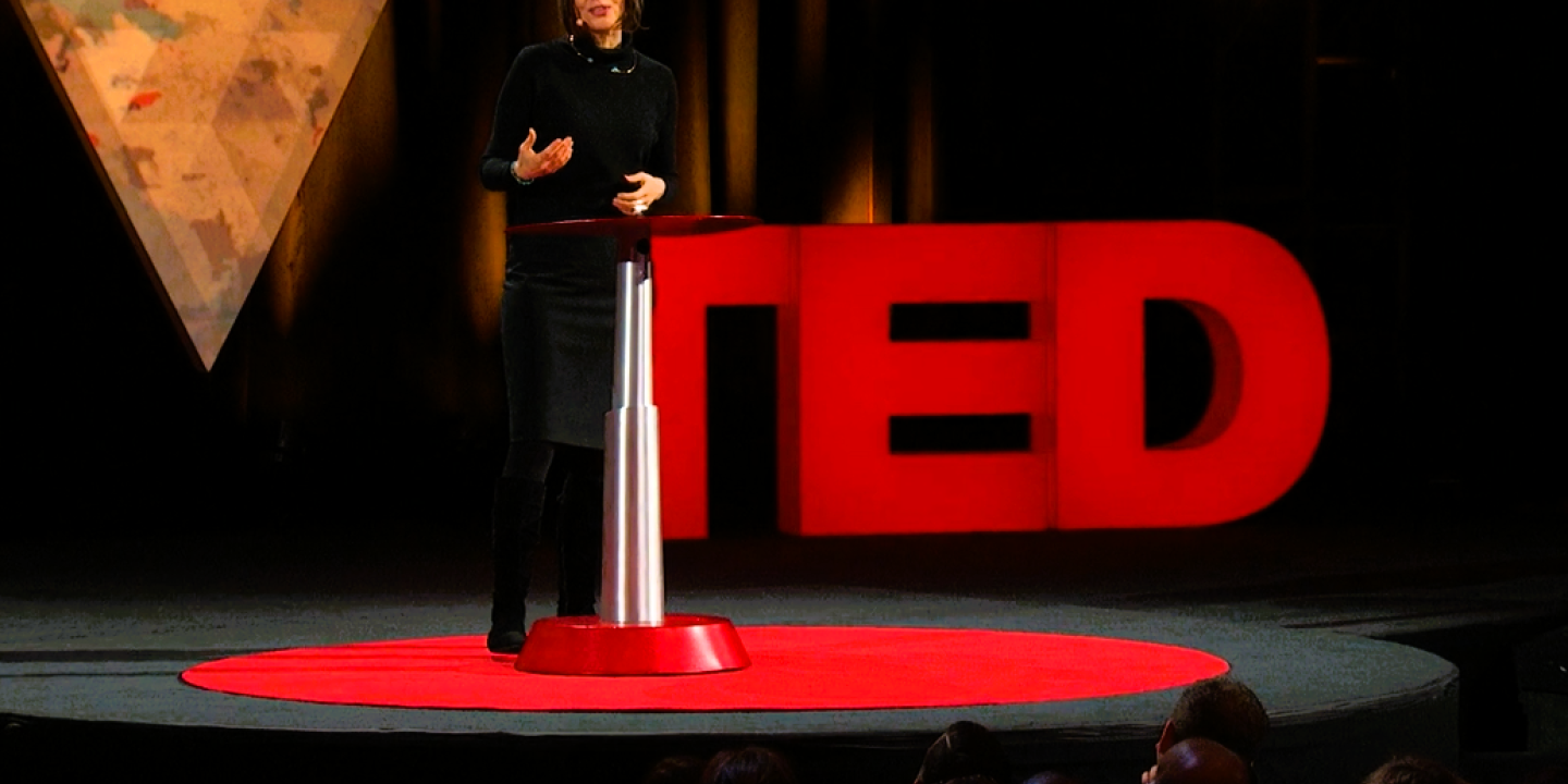 A speaker stands beside a podium in front of a TED sign.