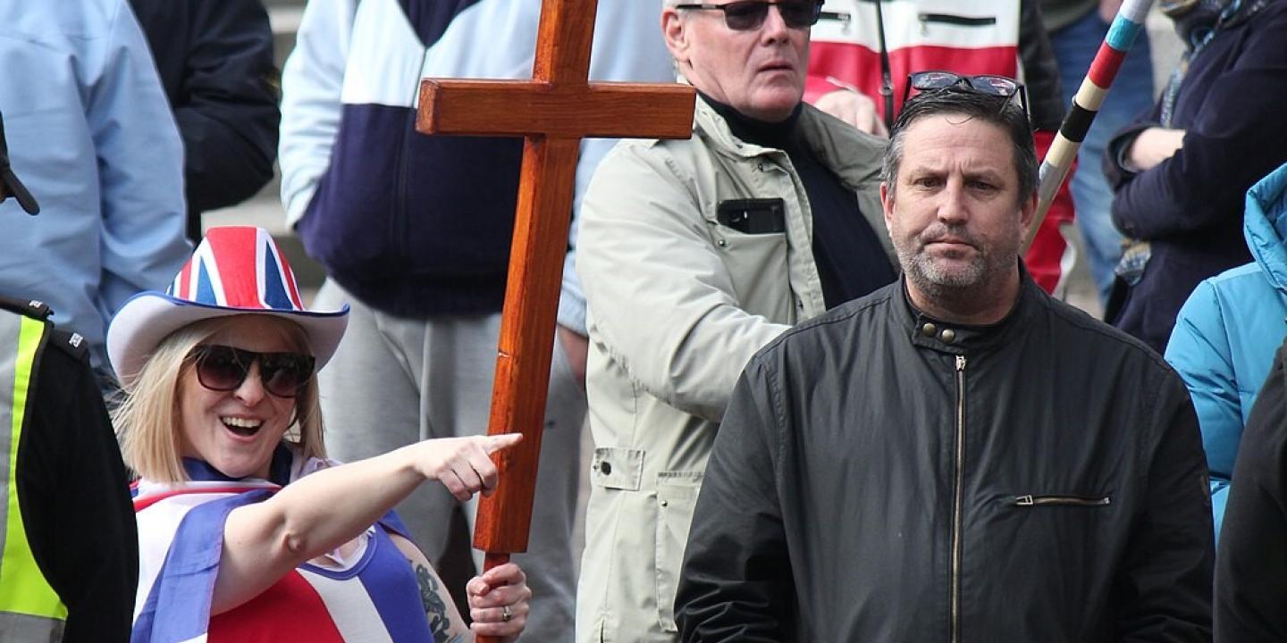 A protester wearing a Union Jack flag and hat and holding a cross, points while a man looks on.