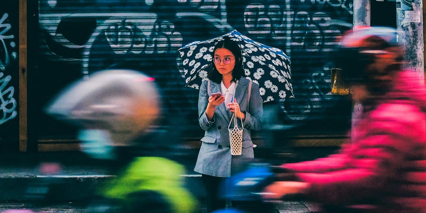 A woman stands across a busy roads, looking up from her phone in a sad way.