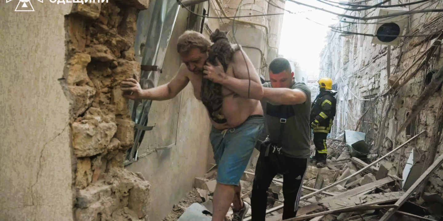 Amid a bombed alley, a victim is helped to walk by a rescue worker