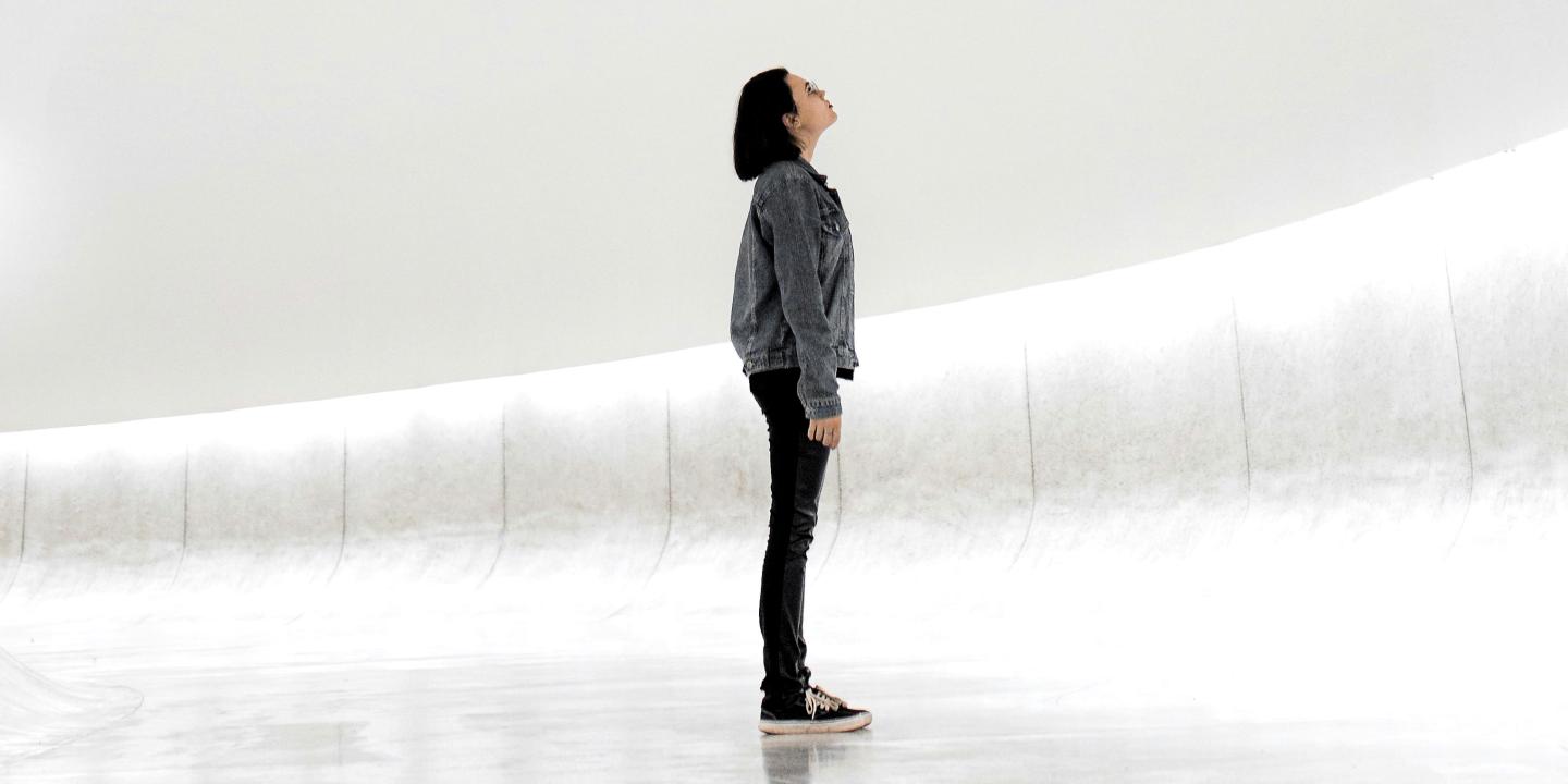A woman stands against a sparse white background looking up.