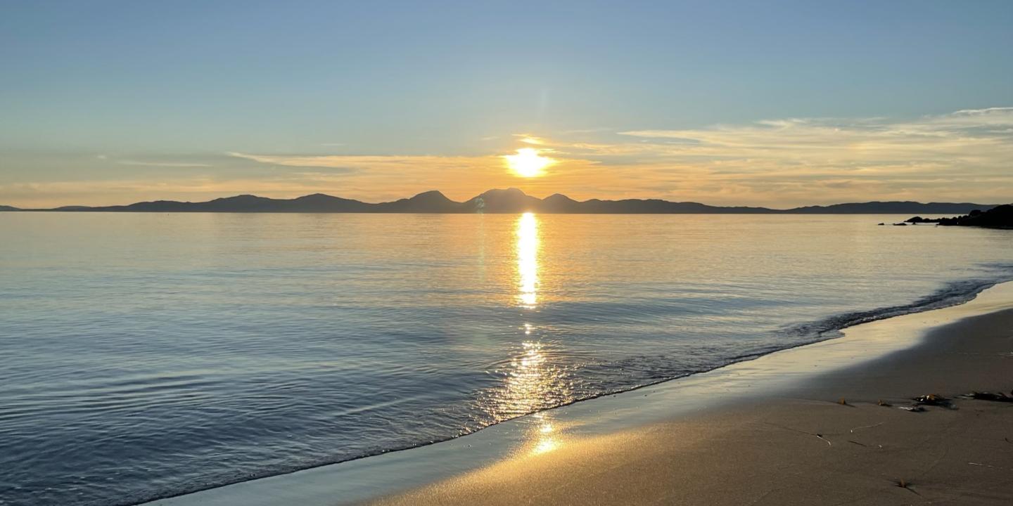 A sunset over an island casts golden light on the sea and a beach.