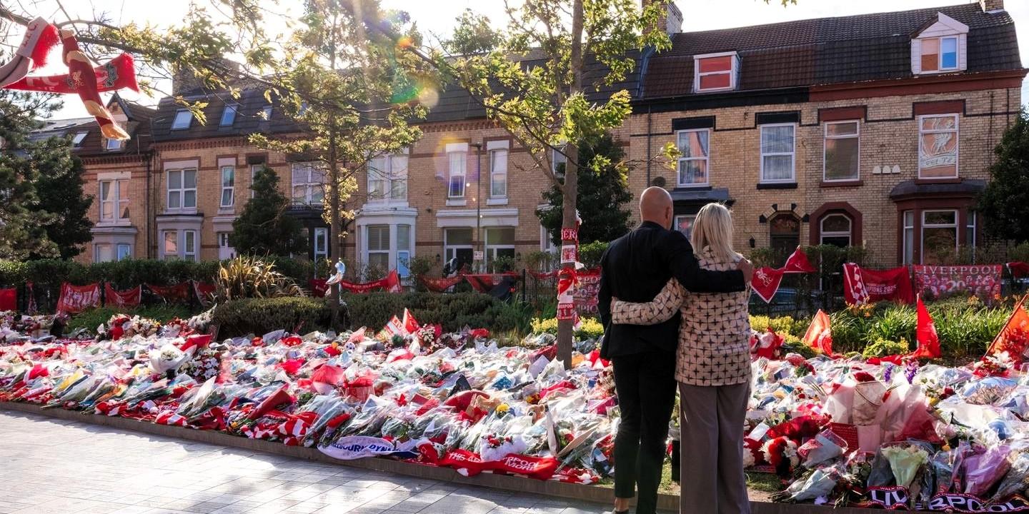 A couple hold each other as they look at floral tributes on the ground