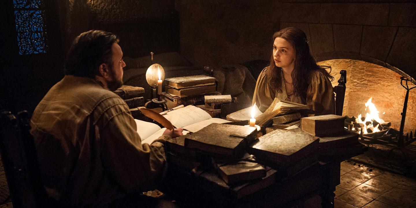 Two people sit at a table strewen with old books lit by candle light.