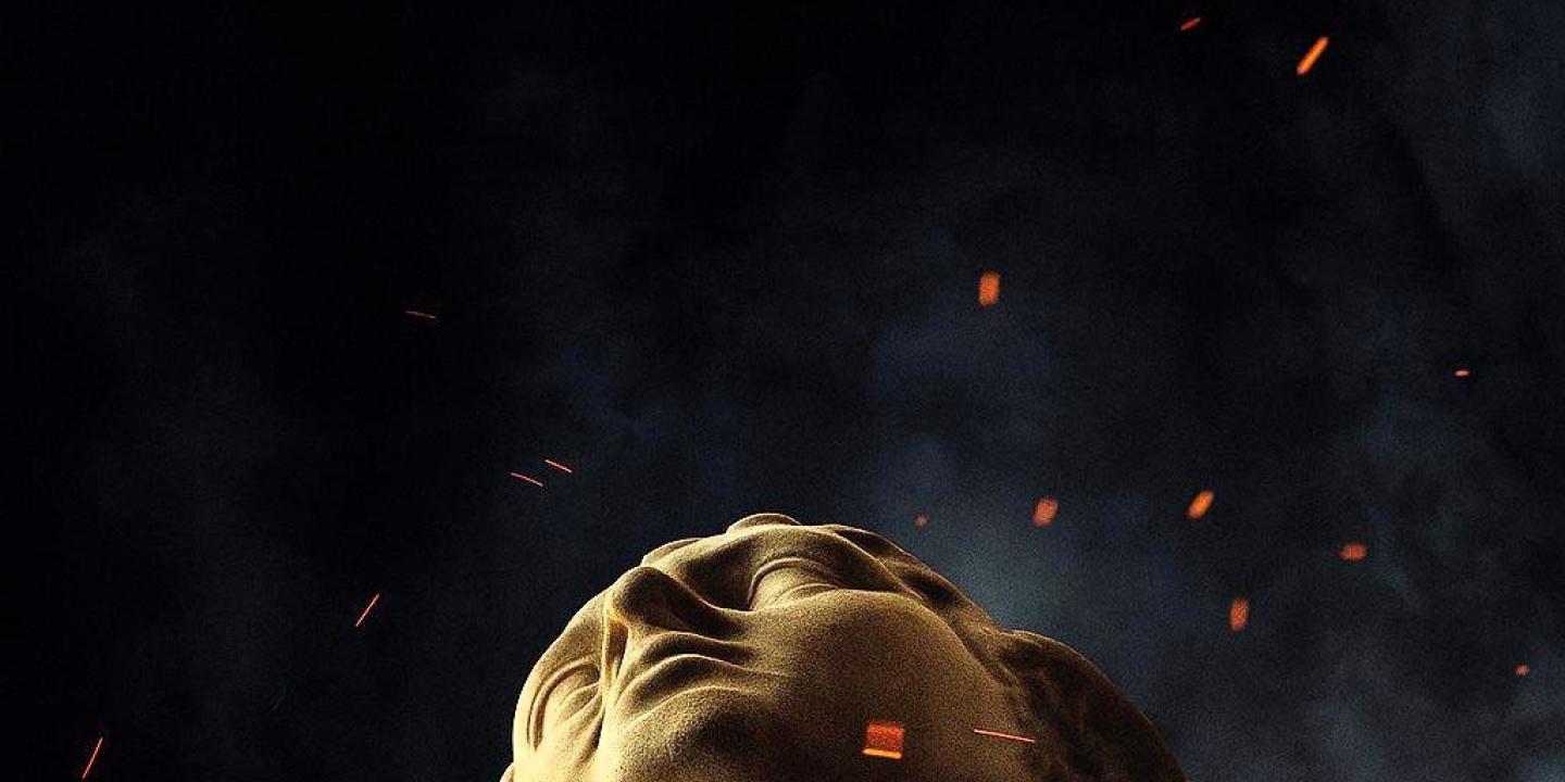 the head of a classical statue looks up amid embers around it.