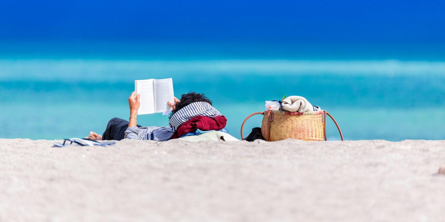 A person lying on a beach holds a book up to read.
