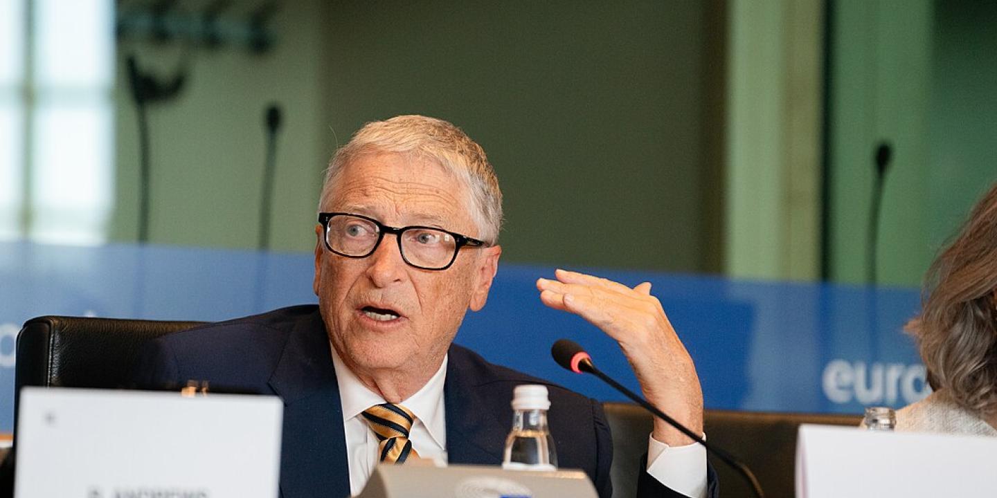 Bill Gates talks from behind a table with a small sign bearing his name.