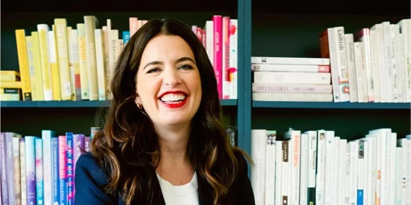 A woman sits and smiles in front of her bookcase.