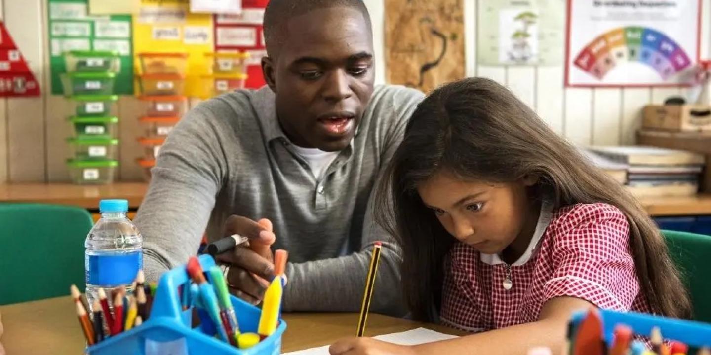 A teacher looks on as a young child concentrates on writing.