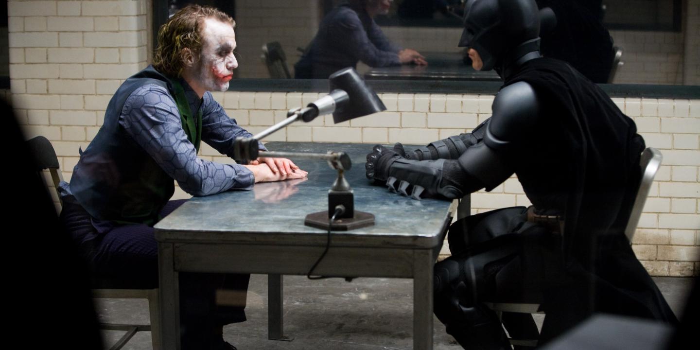 The Joker and Batman sit opposite each other in an interrogation room.