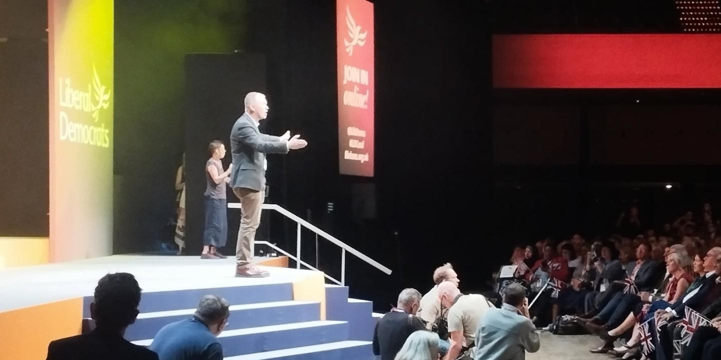 Tim Farron stands on a conference stage with arms wide