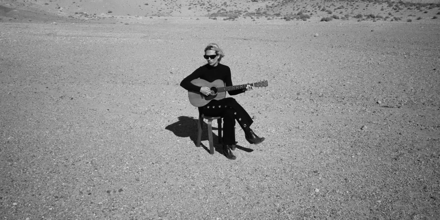 A musician wearing black sits on a chair in a desert holding her guitar.