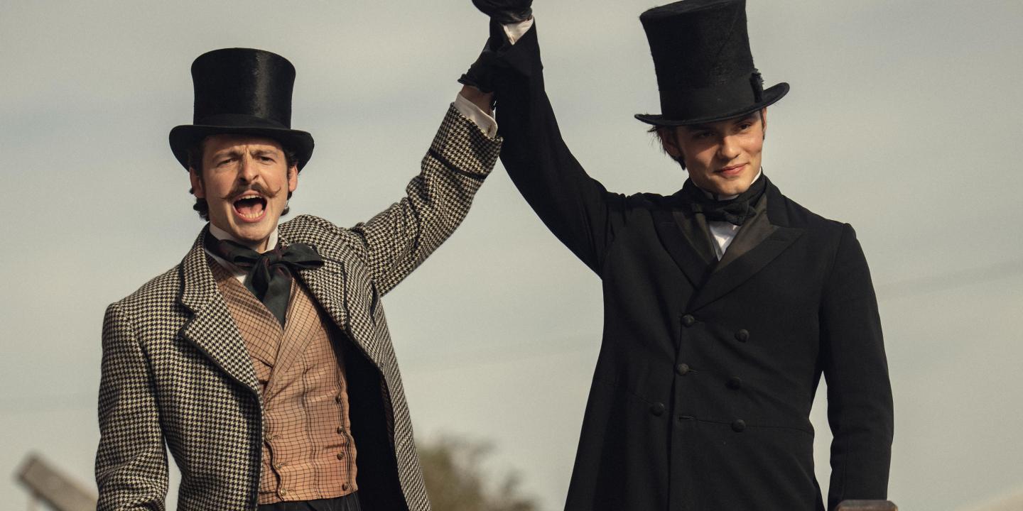 Dressed in Victorian clothes, two brothers raise their arms together.