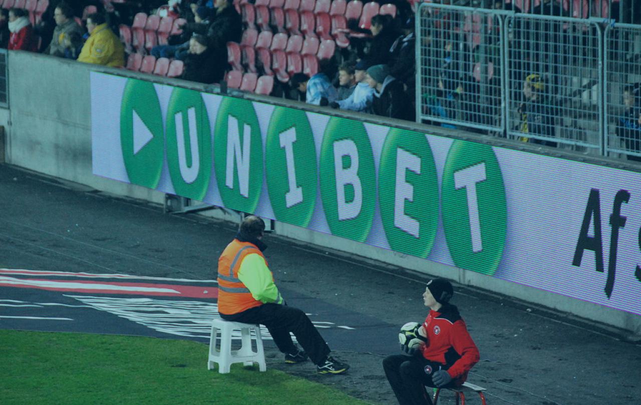 The edge of a football pitch showing an advertising hoarding with a betting brand name on it.