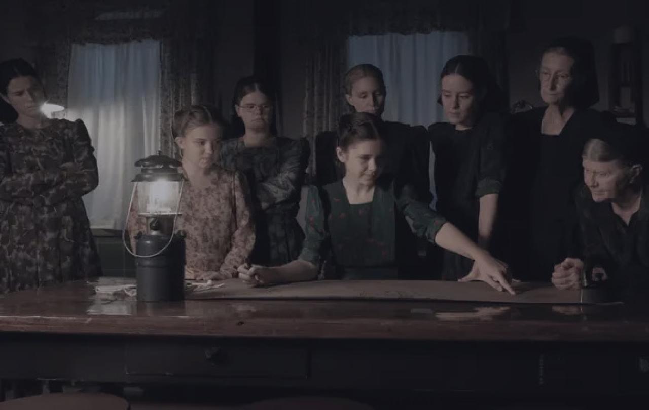 a group of women stand and sit around a table lit by a gas lamp.