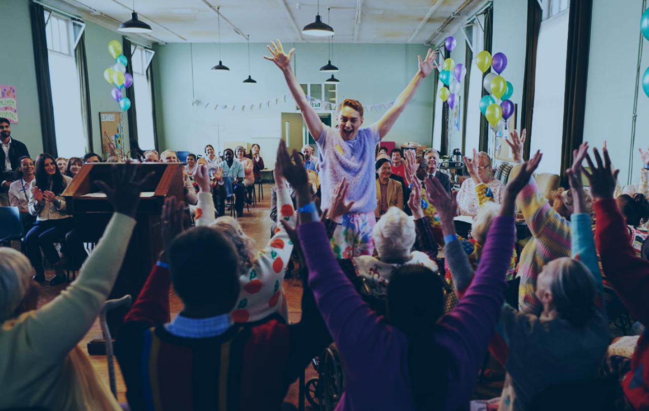 In a hall decorated for a celebration a person stands in front of a seated group, all have their arms raised in celebration.