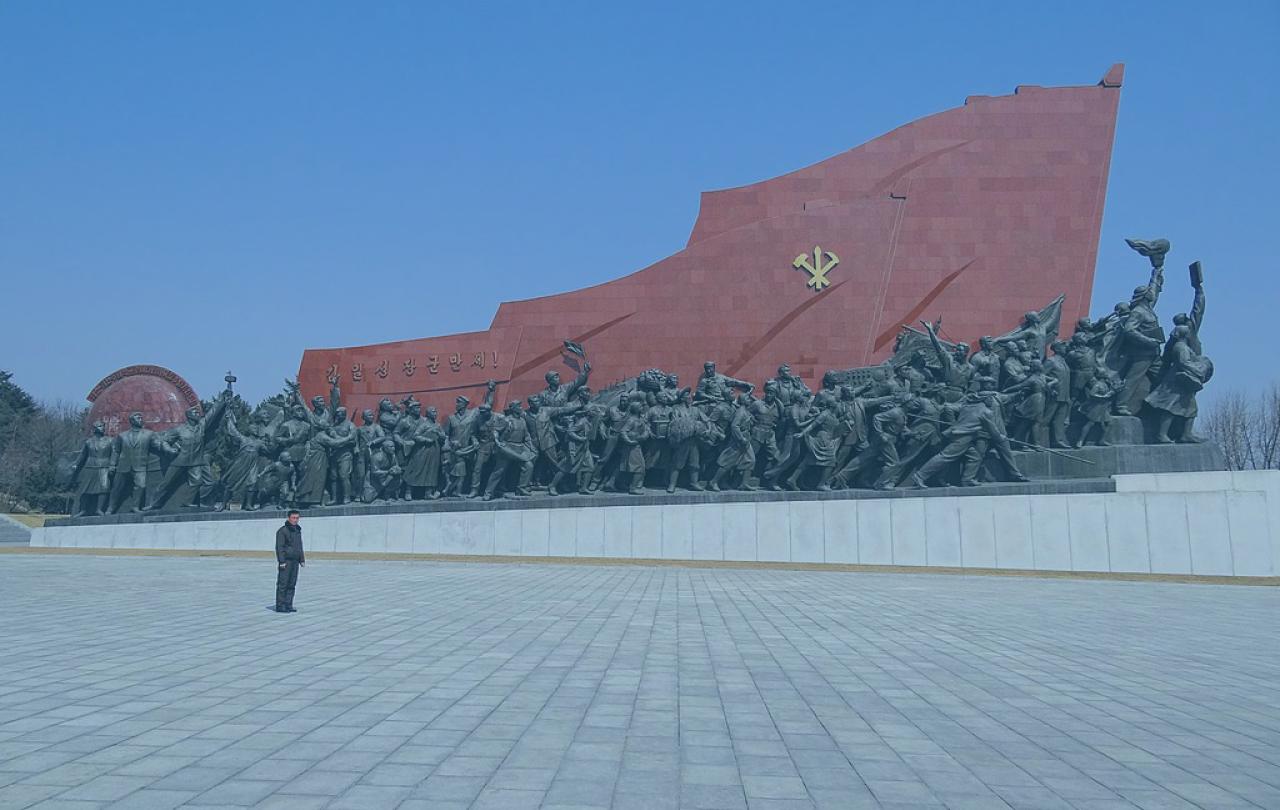 A huge communist monument consists of a red flag wall rising from left to right over a column of statues.