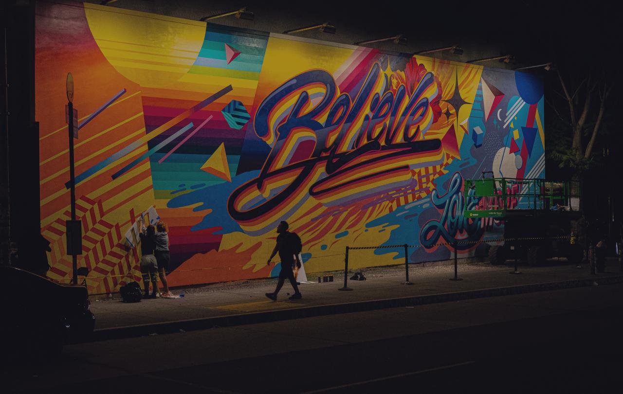 A person walks past a multi-coloured wall of graffiti with the word 'believe' in the centre of it.