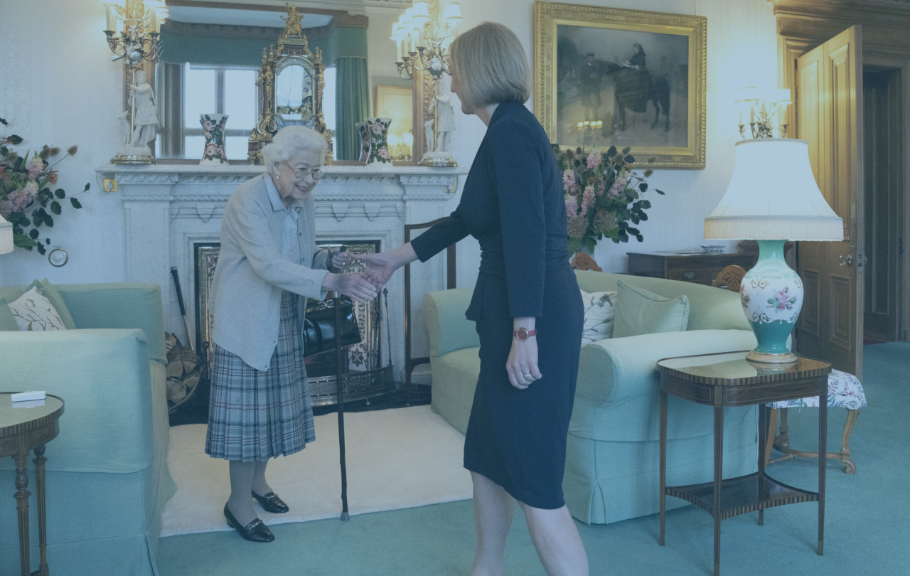 A frail old lady, the late Queen, rises from a sofa to shake hands with an approaching woman.