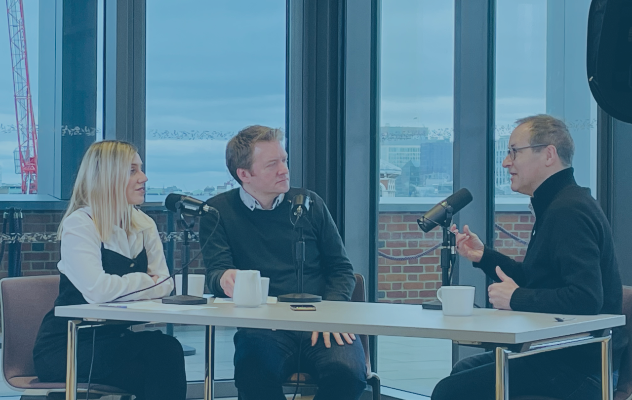 Three people sit around a table recording in front of microphones, one gestures with their hand