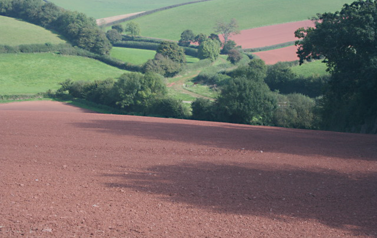A ploughed field of red soil is in the foreground, sloping down into a valley with a track and green fields beyond