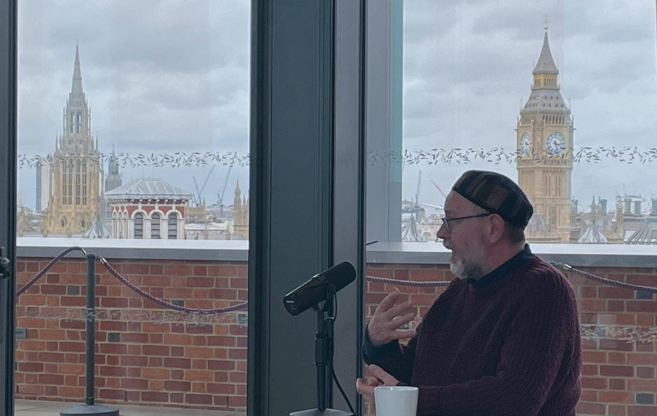 A man sits and speaks into a microphone beside him on a table, while gesturing with his hand. Behind, Big Ben is on the skyline.