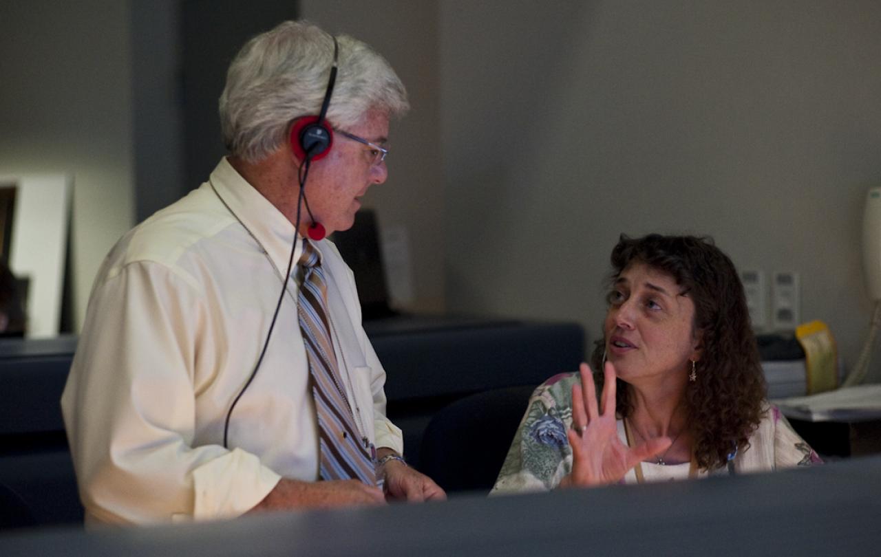 Two scientists talk to each other at console in a space mission control room.