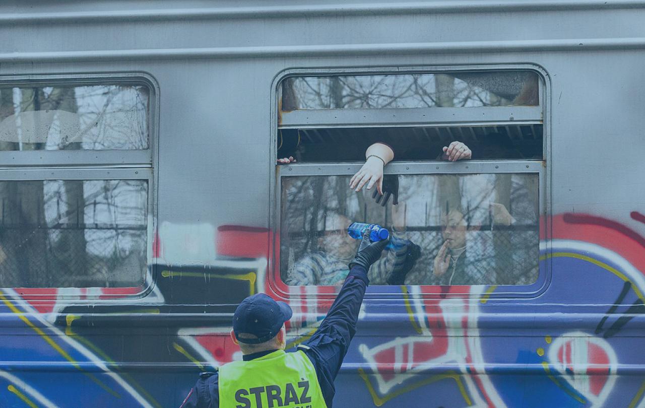 A helper in a yellow vest reaches up to a open train carraige window while offering a bottle. The side of the carraige is covered in graffiti.
