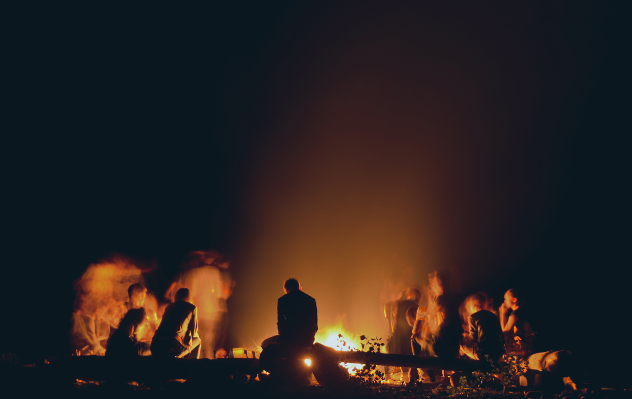 a group of people crowd round a campfire backlighting them in silhouette.
