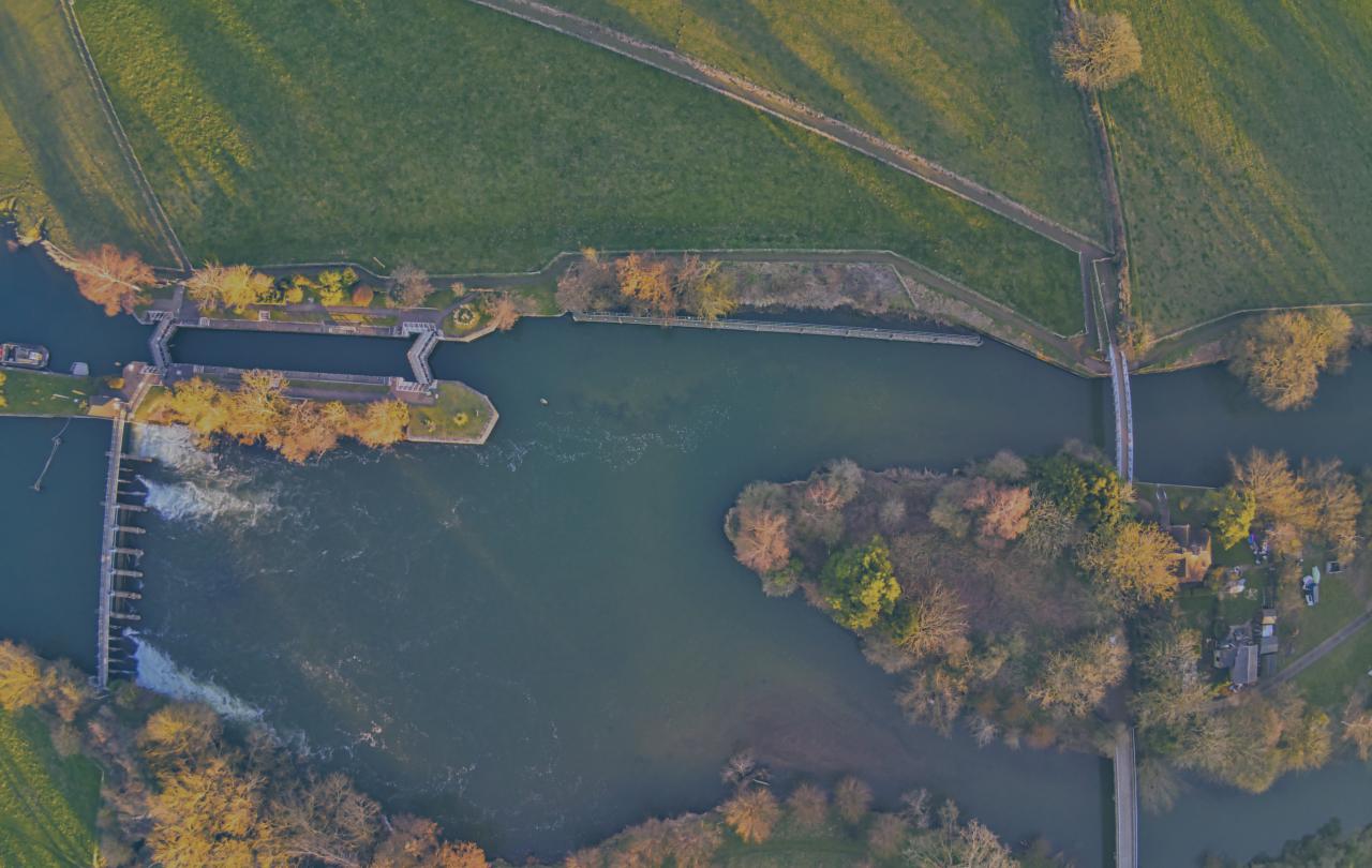 A bird's eye view of a river, with a lock and weir to the left and an island with two bridges to the right.