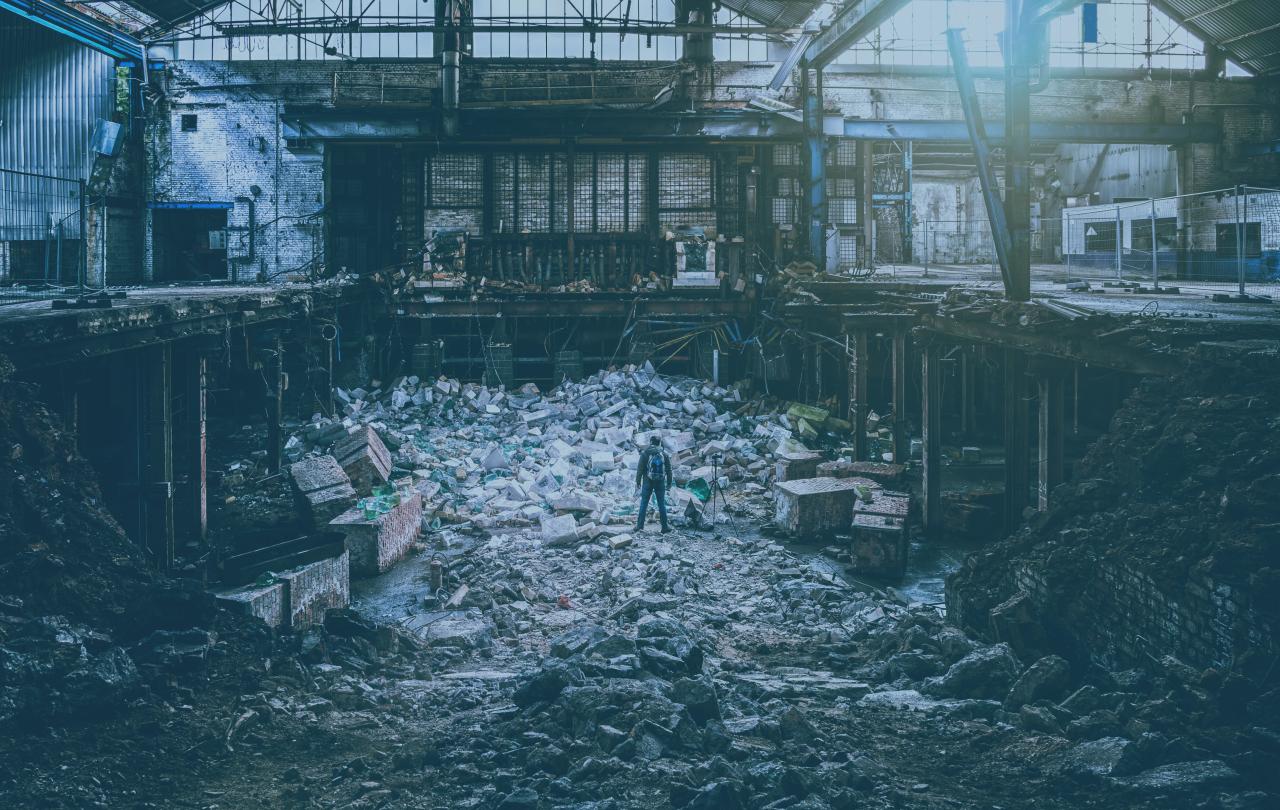 A photographer, standing next to a tripod, atop a pile of rubble is a destroyed factory.