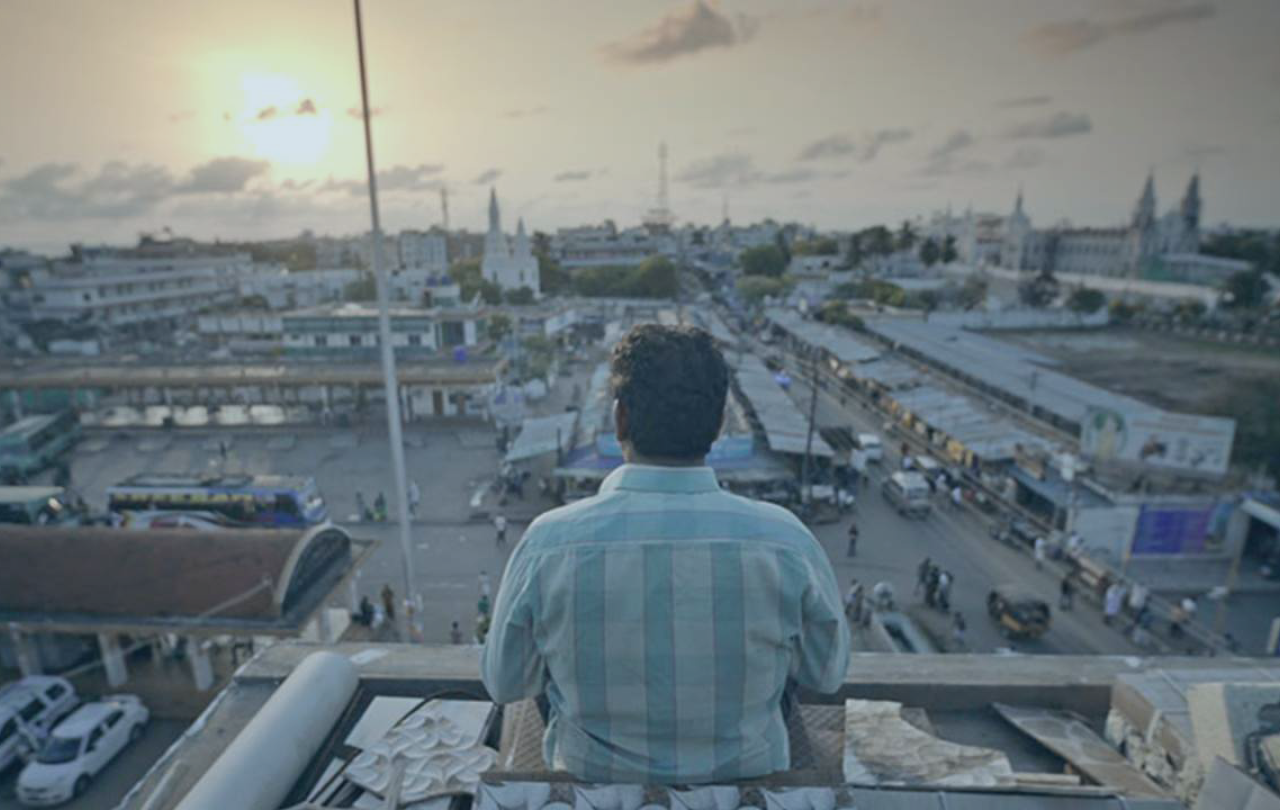 A man sits on the top of a building and looks out over the view of an Indian city as the sun sets.