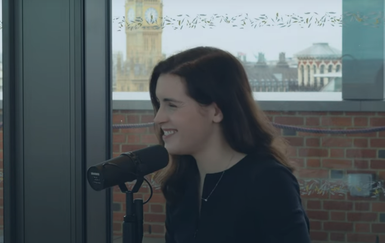 A woman smiles as she speaks into a microphone. In the background is Big Ben.