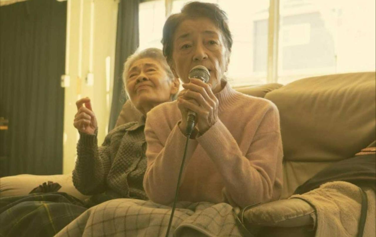 In a retirement home, a older person sings karaoke while the person behind waves a hand.