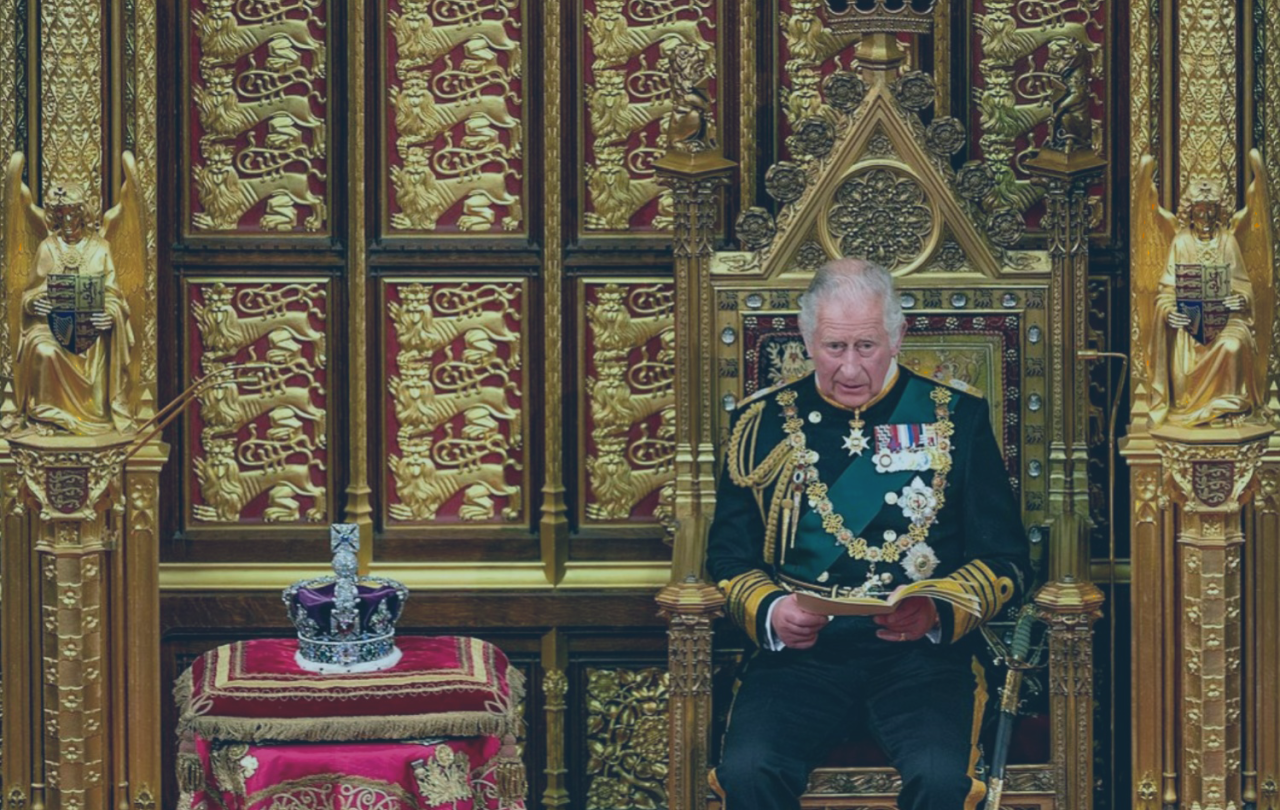 A uniformed Prince Charles sits on a throne reading a speech, beside a crown resting on a cushion